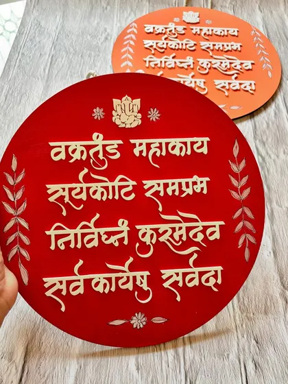 Holding the red 'Vakratunda Mahakaya' wall hanging. This piece is lightweight and easy to hang, instantly adding a spiritual touch to your decor.