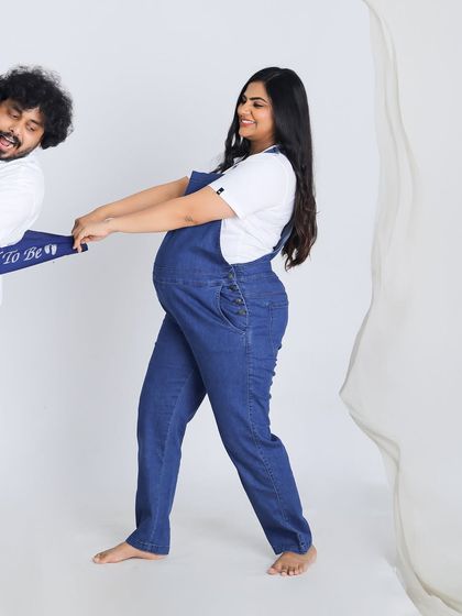 A playful and fun studio shot of an expecting couple. Dressed in casual denim overalls, they share a laugh in this lighthearted and modern maternity photo.