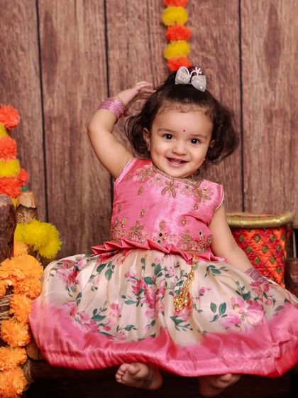 A happy little girl dressed as Radha, surrounded by marigold flowers. Her bright smile and playful pose make this a truly heartwarming photo.
