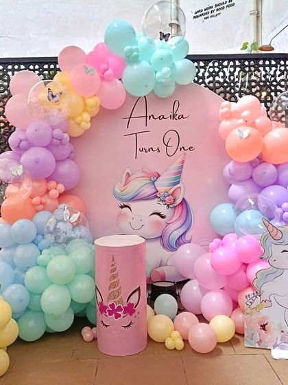 A cheerful and colorful unicorn theme for a first birthday. This outdoor setup features a pink backdrop with a unicorn graphic, a pastel rainbow balloon arch, and unicorn-themed props and standees.