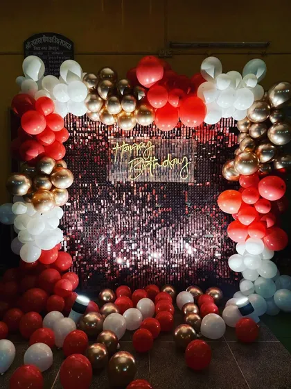 A glamorous birthday photo booth with a red sequin wall. The backdrop is framed by a red, white, and gold balloon arch, creating a perfect spot for party pictures.