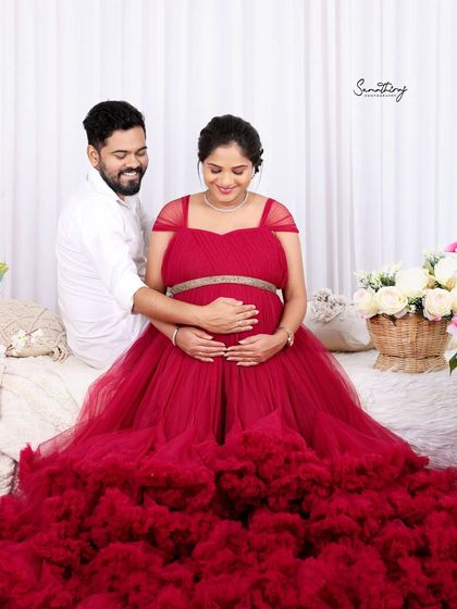 An intimate moment between a couple, both cradling the baby bump. The rich red of the gown and the soft studio lighting create a warm and loving atmosphere.