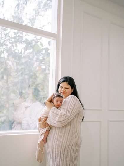 A wider view of the mother and newborn by the window, showing more of the peaceful, light-filled space of the at-home session.