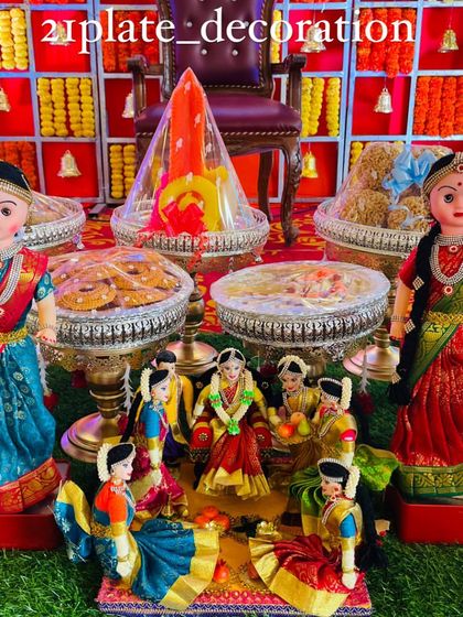 A beautiful and complete baby shower setup with a red and yellow checkered backdrop. The decor includes two large dolls and a central doll scene depicting the Seemantham ceremony.