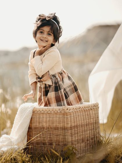 A golden hour portrait of a little girl with a sweet smile, sitting in a wicker basket in a field of tall grass.