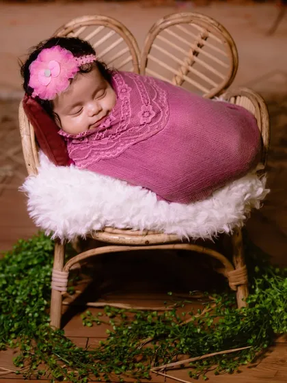 A swaddled newborn sleeping peacefully in a miniature wicker chair, surrounded by greenery for a natural, organic feel.