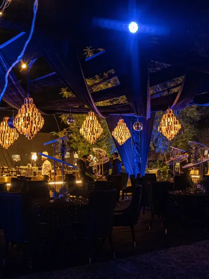A wider view of the outdoor seating area, showing the interplay of the dark draped ceiling, warm chandeliers, and cool blue accent lighting.