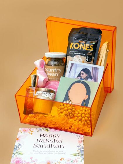 A stylish Rakhi hamper for a sister, presented in a vibrant orange acrylic box. It's filled with coffee, snacks, a scented candle, perfume, and personalized photo items.