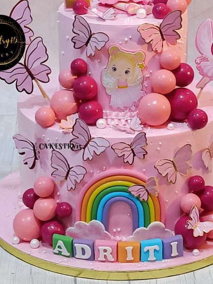 The lower tier of the enchanting 4kg pineapple cake, showing the fondant name blocks, rainbow, and a cascade of pink decorative balls and butterflies.
