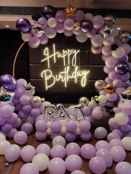A lovely purple-themed balloon ring with the name 'RAYA' in foil letters. The shades of lavender and white, with a neon sign, make for a beautiful party backdrop.