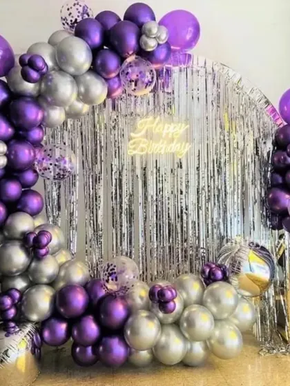 A stylish balloon arch over a silver fringe backdrop. The purple and silver chrome balloons, along with a 'Happy Birthday' neon sign, create a modern and celebratory look.