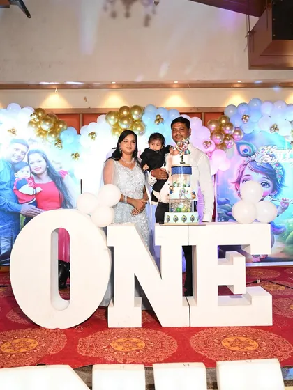 A family poses with their son for his first birthday, standing next to a large "ONE" sign and our Krishna-themed cake.