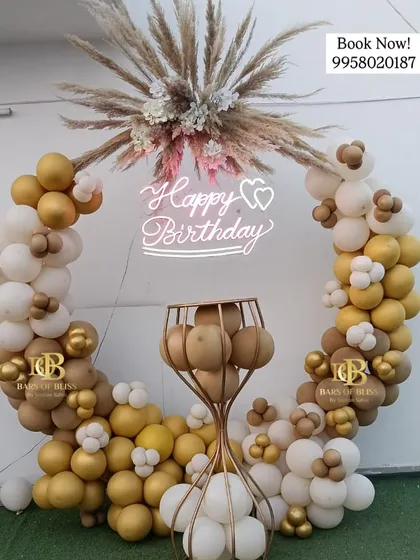 This rustic-chic birthday decor features a full ring of white and gold balloons, topped with a spray of pampas grass. A neon sign and a golden pedestal for the cake complete this elegant setup.