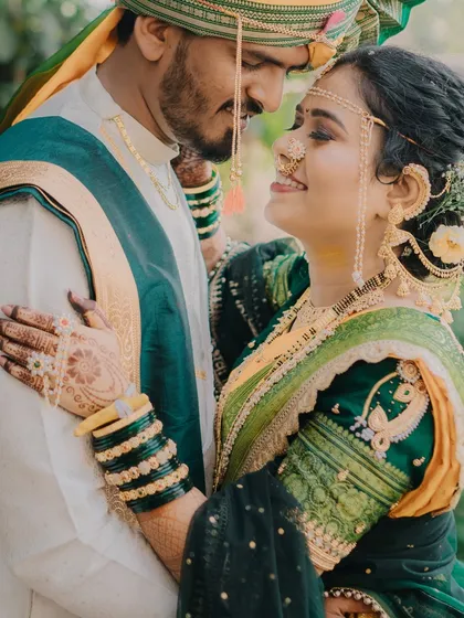 A romantic portrait of a couple in traditional attire, surrounded by nature. The bride's hennaed hand gently resting on her groom's chest shows their closeness and affection.