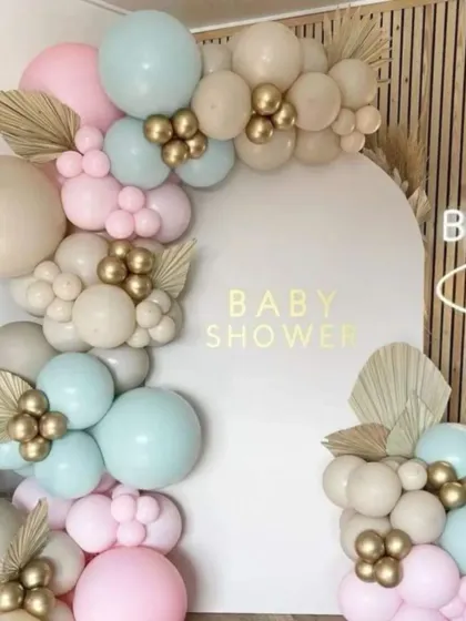 A boho-chic baby shower setup. The pastel blue, pink, and beige balloons are accented with gold clusters and dried palm leaves for a trendy and stylish look.