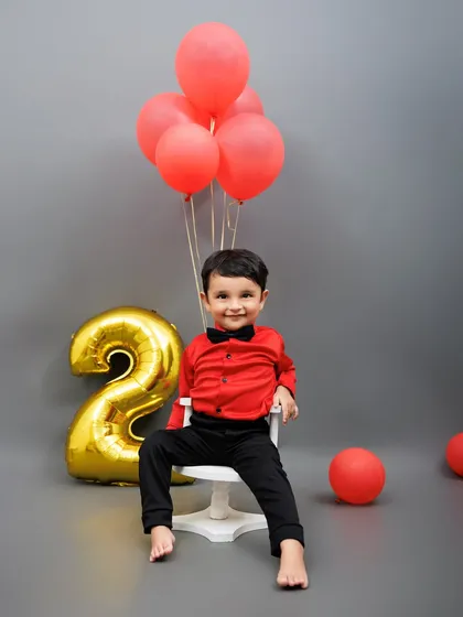 A dapper little boy in a red shirt and bow tie smiles for the camera during his second birthday photoshoot.