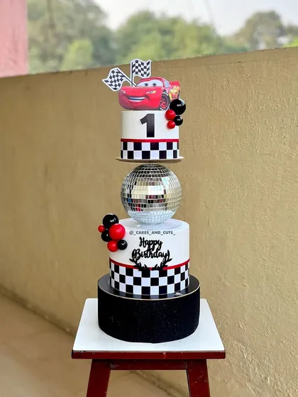 A stunning three-tier Lightning McQueen cake for a first birthday. The design includes a checkered pattern, a disco ball separator, and a fun character topper.