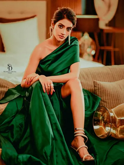 Actress Nusrat Jahan looking elegant in a one-shoulder green satin gown. This full-length shot on a sofa showcases the dramatic silhouette of the dress and her poised, high-fashion pose.