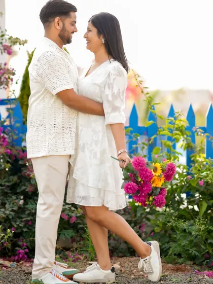A sweet, candid moment with the couple in matching white outfits against a vibrant floral backdrop. Their easy smiles and relaxed posture show the comfort and joy they share.