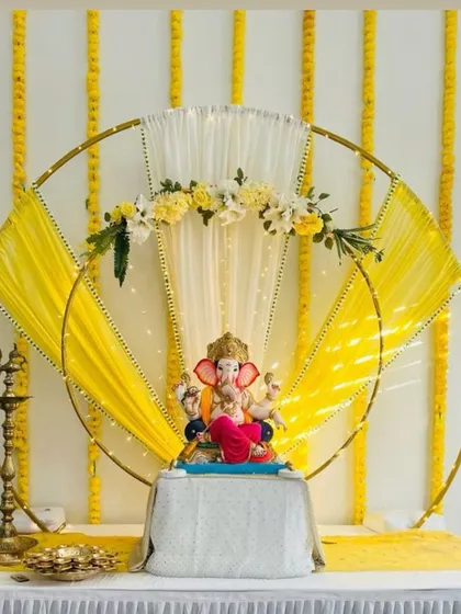 A modern and artistic Ganpati setup featuring two overlapping golden rings draped with yellow and white fabric. The backdrop of hanging marigold strings adds a traditional touch to this contemporary design.