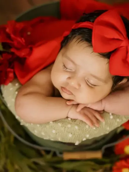 A beautiful two-month-old baby girl sleeping peacefully in a bucket prop. The vibrant red bow and flowers add a lovely pop of color to this newborn portrait.