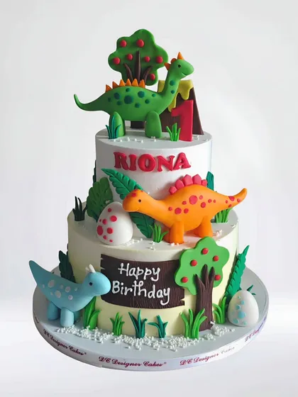 A delightful two-tier dinosaur-themed cake for a first birthday. This whipped cream cake is decorated with friendly, colorful fondant dinosaurs, trees, and a volcano for a fun prehistoric party.
