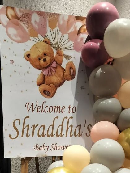 A close-up of a welcome sign for 'Shraddha's Baby Shower'. The design features a cute teddy bear holding onto balloons, matching the party's theme.