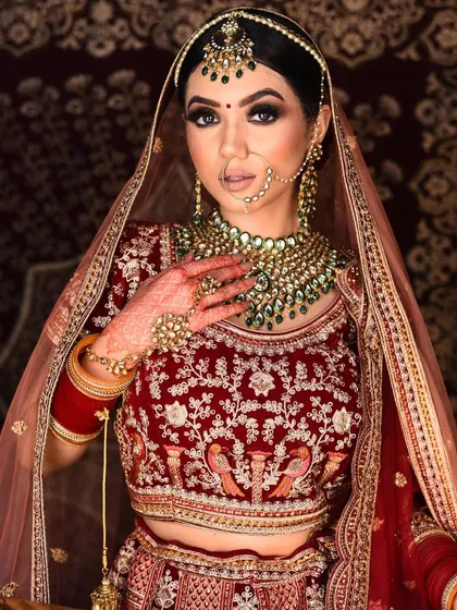 A classic bridal pose in a red velvet lehenga with peacock motifs. The green-beaded Kundan jewellery provides a beautiful, traditional contrast.