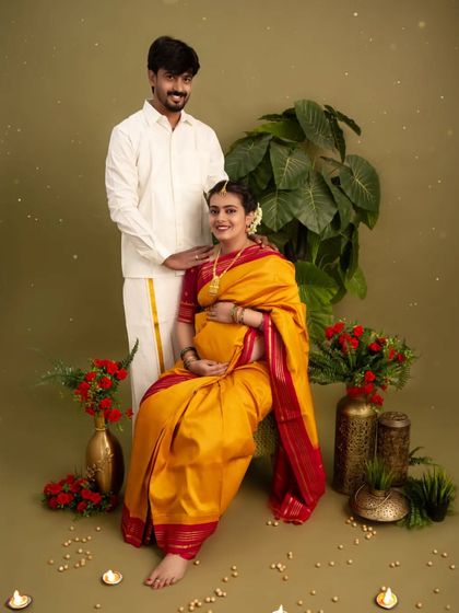 A beautiful couple's portrait showcasing traditional South Indian attire. The husband stands beside his seated wife, both sharing a happy moment in a simple, elegant studio setup.