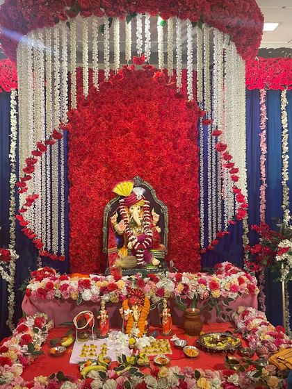 A powerful and traditional Ganpati decoration using a bold red, white, and blue color scheme. The backdrop features a dense red flower panel in the shape of a temple arch, with curtains of white flowers on either side.