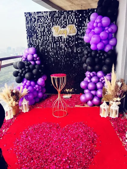 A modern and chic proposal decoration on a balcony. This setup features a black shimmer wall backdrop, bold purple and black balloon arrangements, and a red carpet covered in flower petals for a dramatic effect.