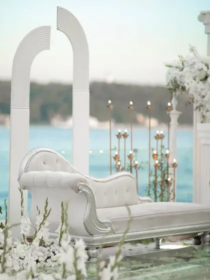 A chic and modern photo seating area set against an ocean backdrop, featuring a white chaise lounge and minimalist white arches.