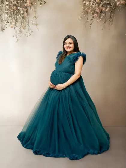 A full-length shot of the mom-to-be in a gorgeous teal gown, set against a simple, elegant backdrop with hanging flowers. Her happy expression and the beautiful dress are the focus of this portrait.