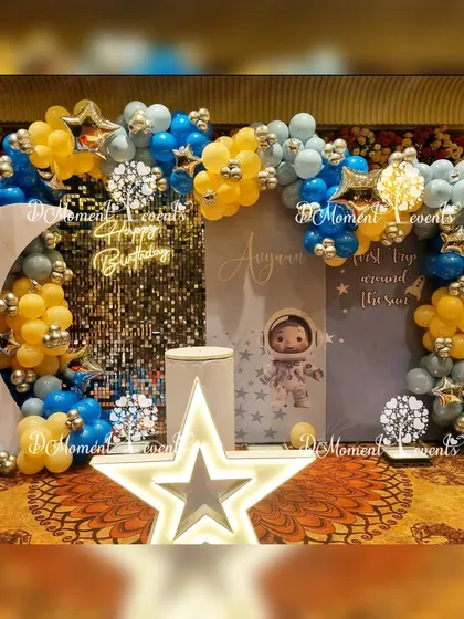 An out-of-this-world space-themed stage for Ayvaan's first trip around the sun. The decor features a moon cutout, a light-up star, an astronaut prop, and a blue and yellow balloon arch against a sparkling sequin backdrop.