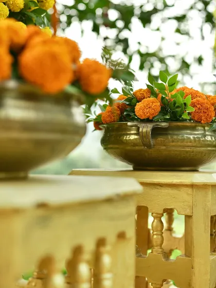 A pair of traditional brass bowls filled with orange marigolds, set on ornate golden stools. This arrangement is a perfect example of classic props used in Haldi and wedding decor.