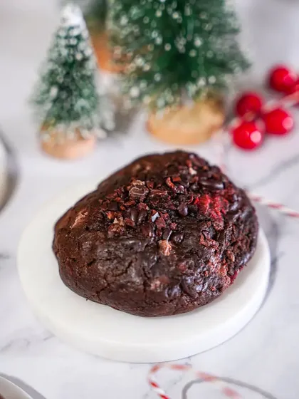 A decadent black forest cookie, shot on a clean marble surface with festive props. The focus is on the rich chocolate and cherry textures of this gourmet Christmas treat.