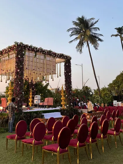 An elegant outdoor mandap setup at dusk. The design features a canopy of woven details and floral chandeliers, with rich velvet seating for a touch of royalty against the open lawn.