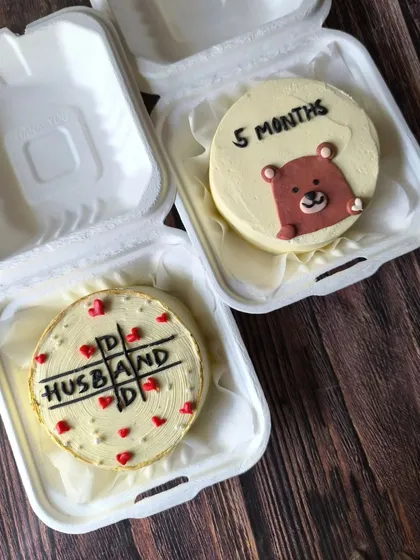 A sweet set of matching bento cakes for a father and son. One cake marks a "5 months" milestone with a cute bear, while the other is a "Dad Husband" tic-tac-toe design for the father's birthday.
