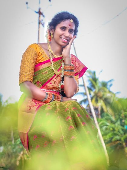 An artistic outdoor shot using foreground blur. The green leaves in the front create a soft frame, drawing the viewer's eye to the subject's gentle smile and traditional attire.