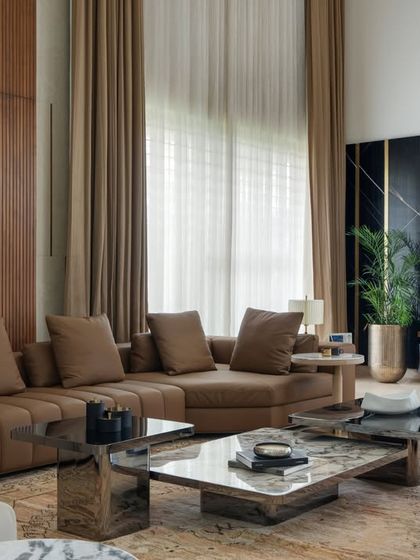 The double-height living area in the Celadon project features a striking black marble wall with brass inlays for the media unit, contrasting with the warm wood paneling. The large sectional sofa and layered coffee tables create a comfortable yet sophisticated space for family gatherings.