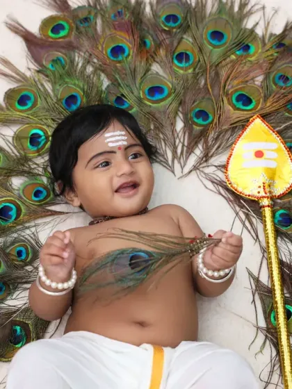 Another adorable shot of the baby as Lord Murugan, smiling and holding a single peacock feather. The simple white background keeps the focus on the baby and the symbolic props.