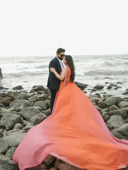 A dramatic shot on a rocky Goa beach, where the bride's vibrant orange gown flows in the wind against the powerful waves, creating a moment of pure cinematic romance.