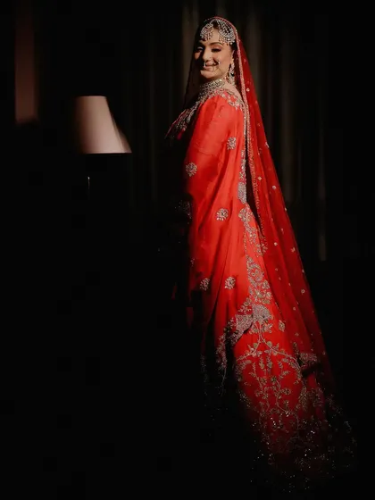 A dramatic bridal portrait using low-key lighting. The single light source illuminates the bride's smiling face and the rich red of her outfit, creating a mysterious and elegant mood.