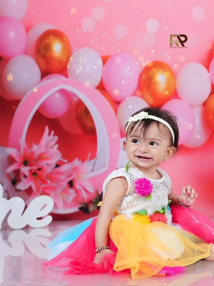 A little girl in a rainbow dress sits beside her cake during her first birthday photoshoot. The pink balloon backdrop and floral props create a bright and cheerful setting.
