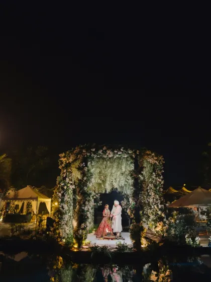 A stunning wide shot of the nighttime ceremony, the floral mandap glowing under the dark sky, its reflection shimmering in the water.
