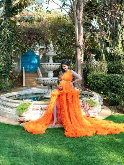 A beautiful, wide-angle shot of a mom-to-be in a flowing orange gown, posing by a garden fountain. The entire scene feels grand and luxurious.