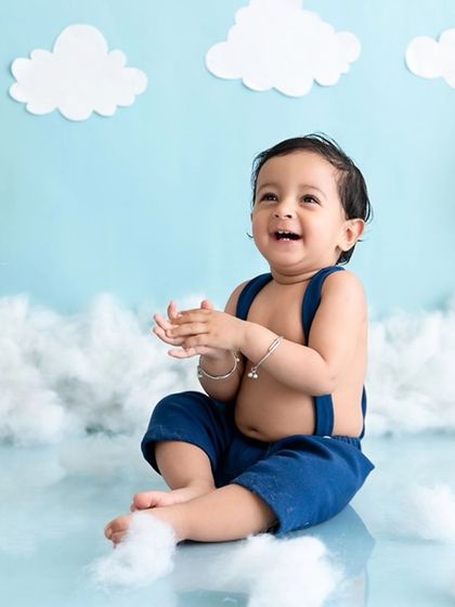 A happy baby sits amidst a sky-themed backdrop with fluffy clouds and balloons, clapping his hands with joy.