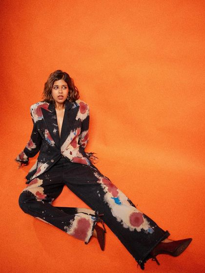 A dynamic, full-body shot of Sejal Kumar against a vibrant orange background. The seated pose is unconventional and full of energy, perfectly capturing the artistic vibe of the hand-painted Huemn suit.