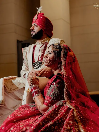 A regal portrait of the couple, with the bride seated, showcasing her elaborate red lehenga and the groom standing beside her.