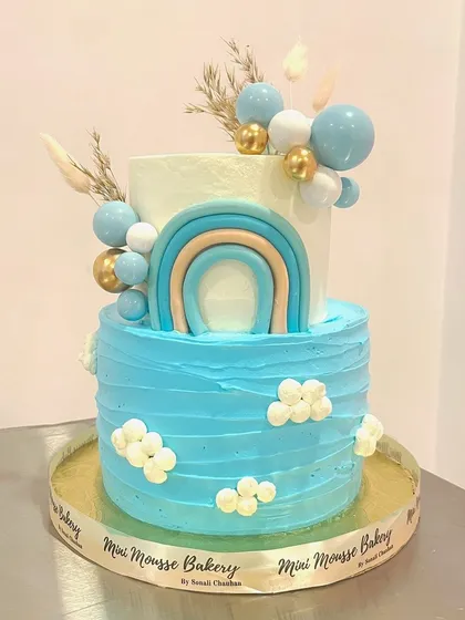 A sweet two-tier cake for a baby boy's naming ceremony. The design has a blue rainbow, fluffy clouds, and a balloon bouquet, all in a lovely pastel blue palette.